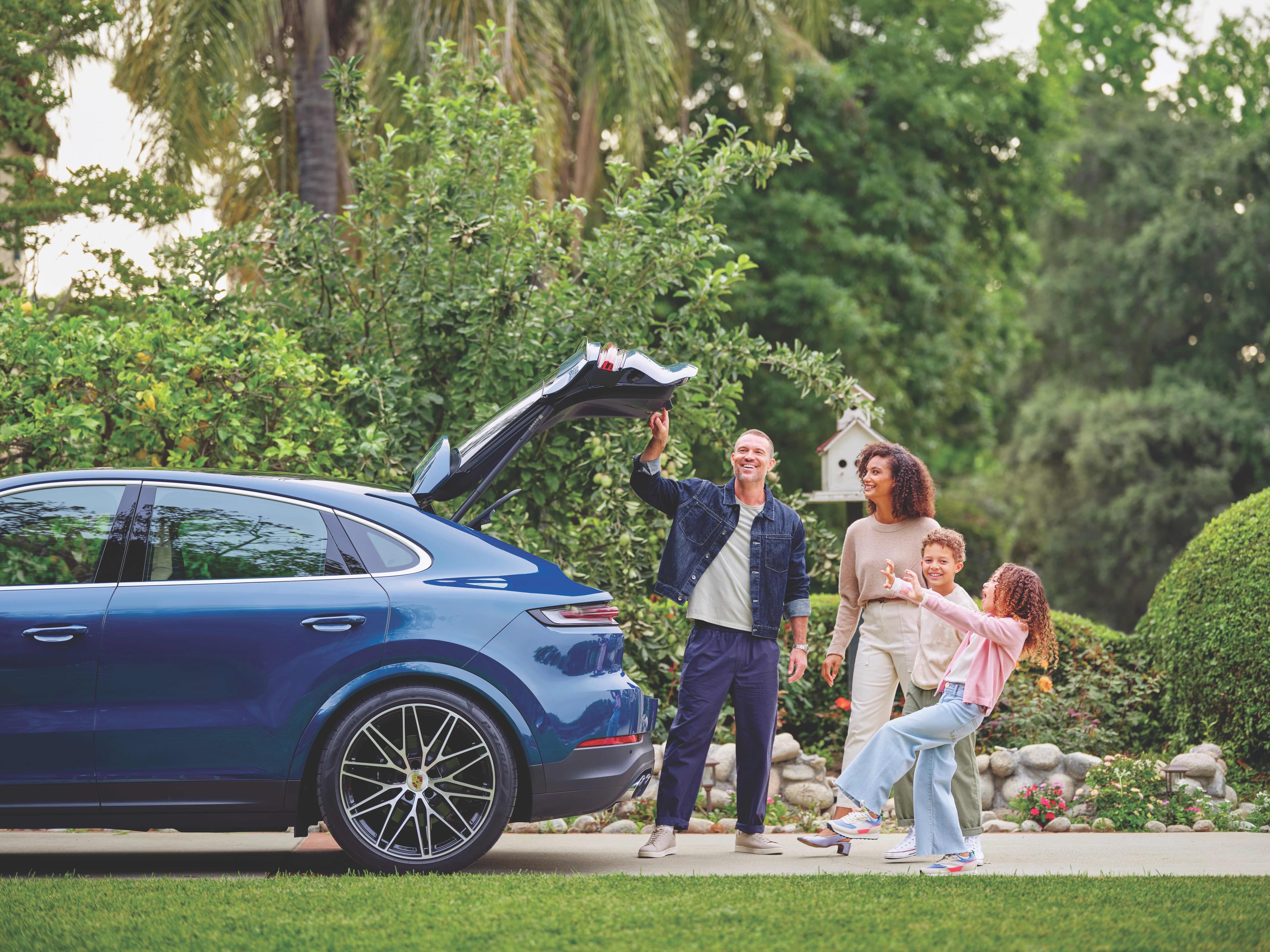 A family of four stands by a blue Porsche Macan SUV with an open trunk, smiling  in a lush garden setting.